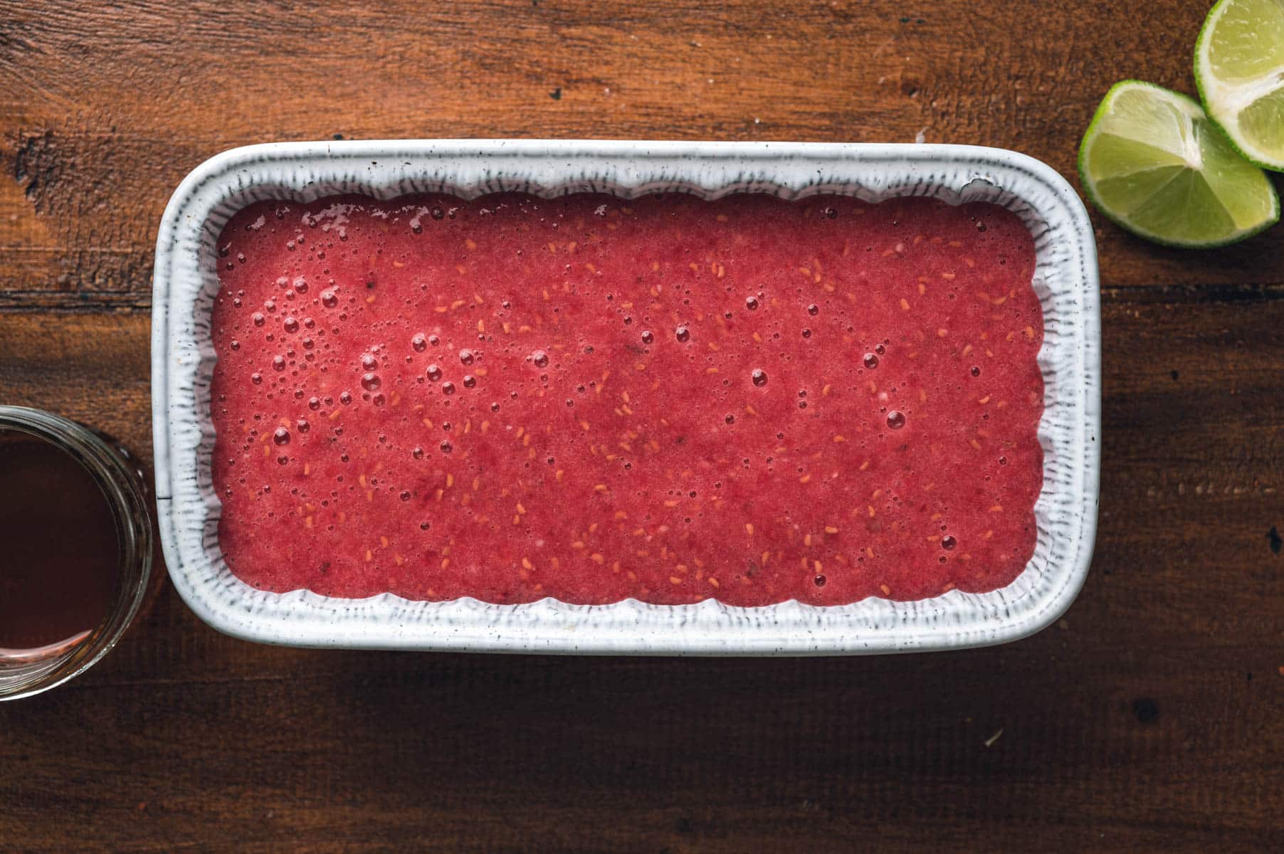 A rectangular dish filled with a red, raspberry sorbet sits on a wooden surface next to a halved lime and a small glass container holding a dark liquid.