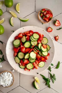 A bowl of sliced cucumbers and strawberries on a tiled surface, surrounded by lime wedges, mint leaves, a bowl of crumbled cheese, and a small bowl of strawberry tops.