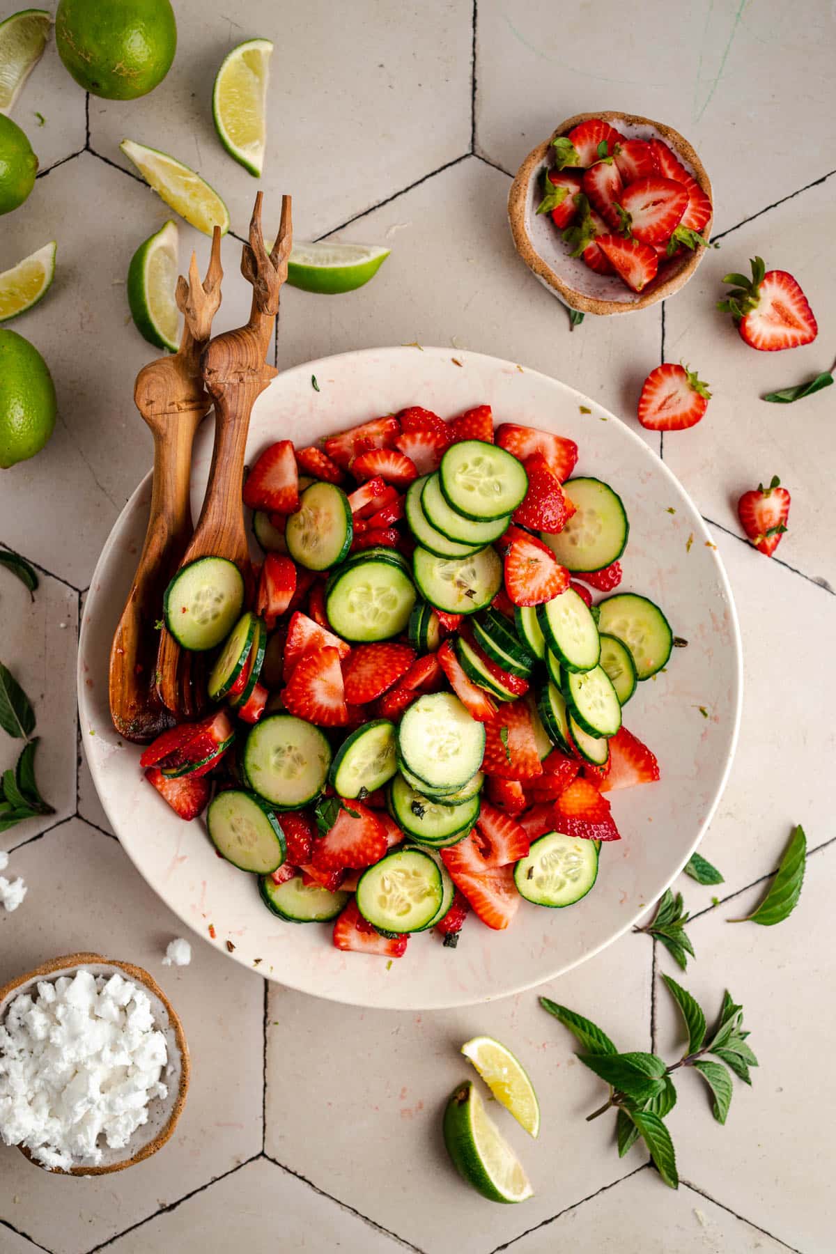 A bowl of sliced cucumbers and strawberries with wooden salad tongs. Surrounding items include partially sliced limes, fresh herbs, feta cheese in a small bowl, and scattered cucumber and strawberry pieces.
