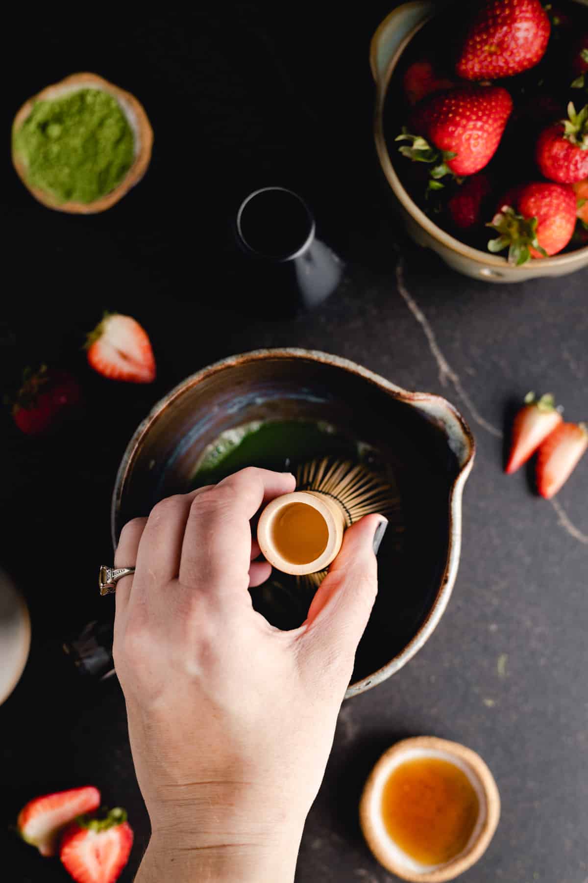 Close-up of a hand holding a small bowl above a larger ceramic bowl containing green powder. Surrounding the bowls are fresh strawberries, a small bowl of green powder, and a black container on a dark surface.