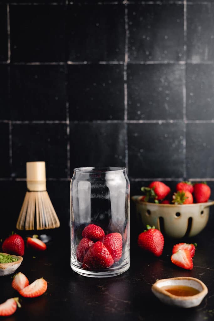 A clear glass with strawberries inside, placed on a dark surface. Surrounding items include more strawberries in a bowl, a small whisk, and a cup, against a dark, tiled background.