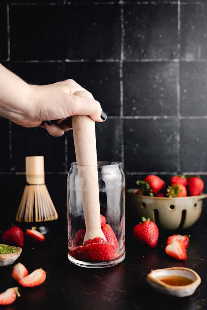 A hand uses a wooden muddler to crush strawberries in a glass. Nearby are whole strawberries in a bowl, a whisk, and a small dish of green powder, all set against a dark tiled background.