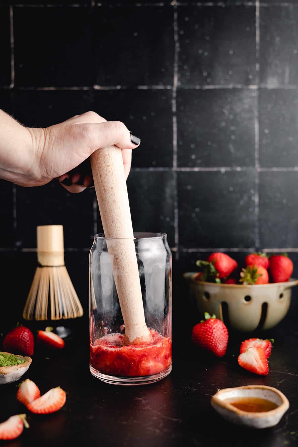 A hand using a wooden pestle to mash strawberries in a glass jar. Fresh strawberries and a small bowl with a liquid are on the table, with a whisk and a container of strawberries in the background.