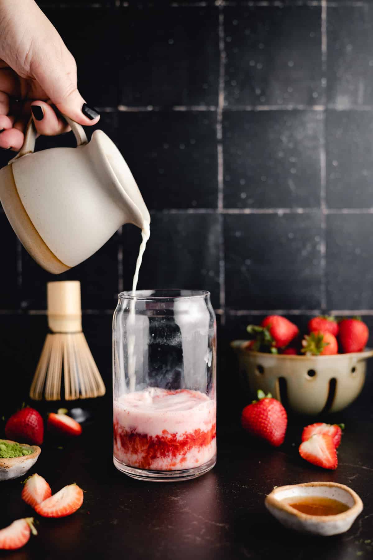 A hand pours milk from a small ceramic jug into a clear glass with a strawberry mixture at the bottom. Fresh strawberries and a ceramic bowl with more strawberries are in the background.