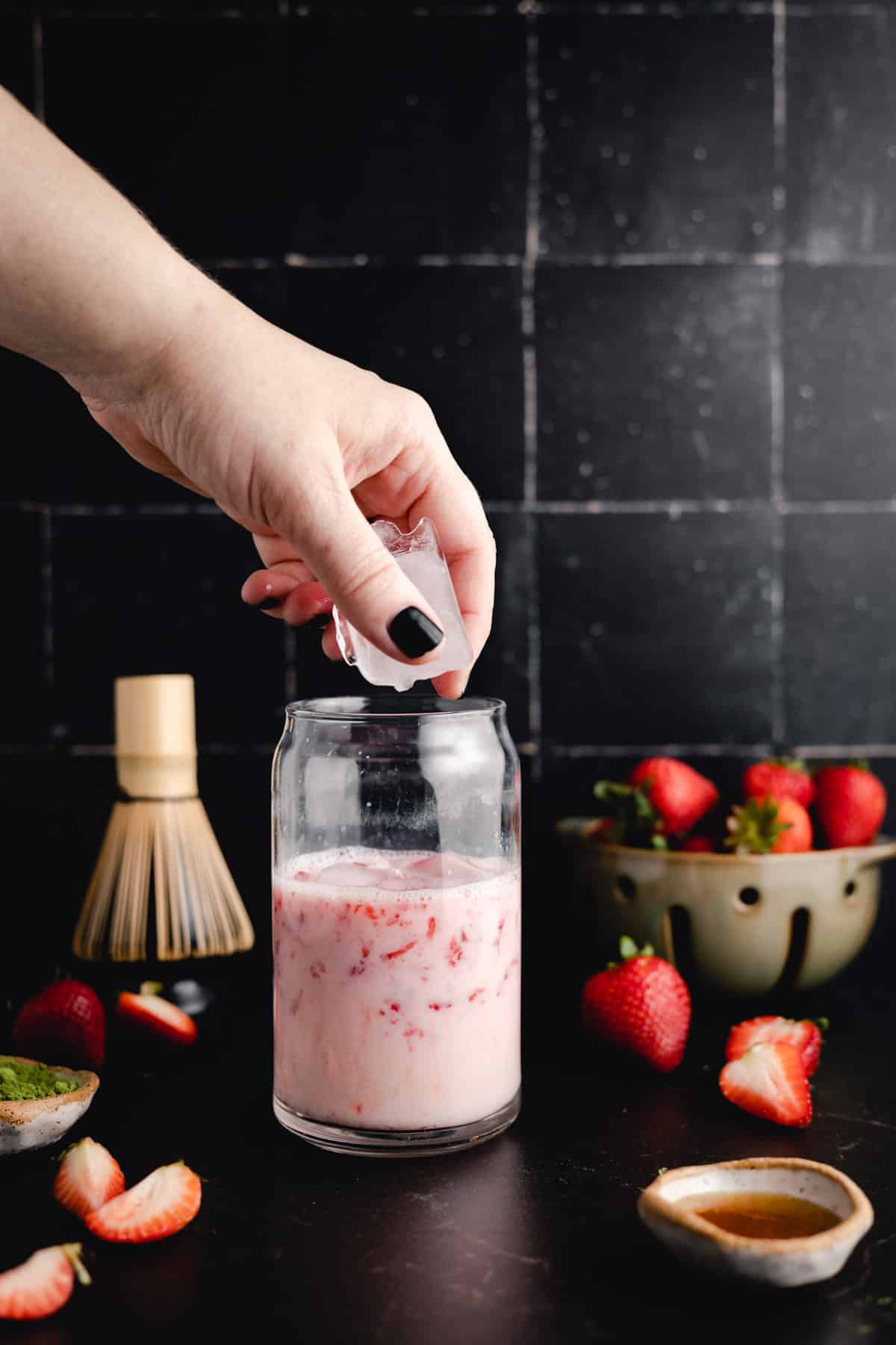 A hand with black nail polish is adding ice to a glass jar of pink strawberry beverage. Fresh strawberries, a small bowl, and a whisk are on the dark surface around the jar.