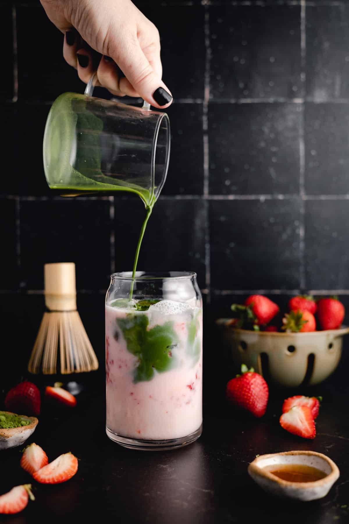 A hand is pouring green matcha into a glass of milk and strawberries. A bowl of strawberries and matcha tools are in the background on a dark countertop.