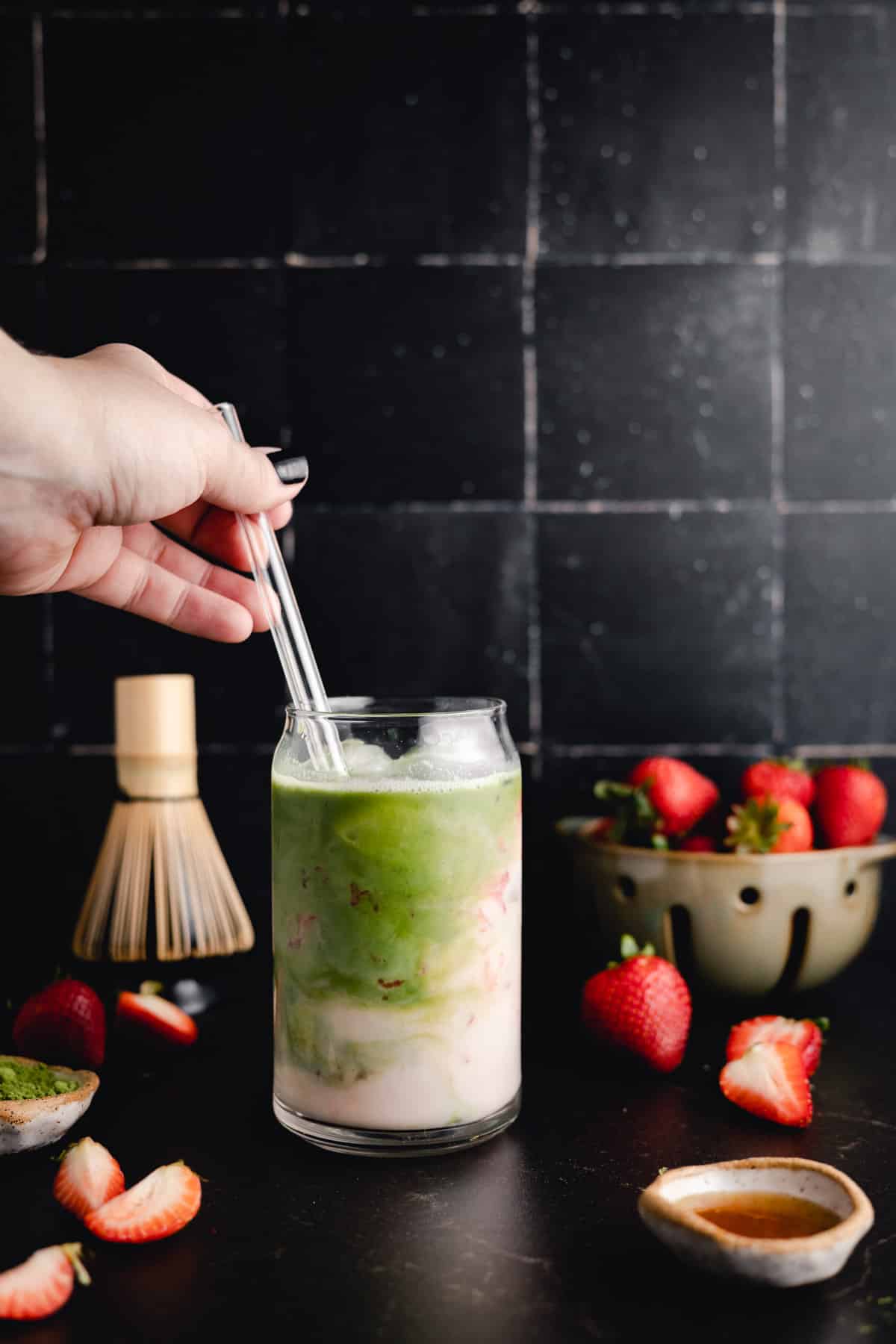 A hand stirs a layered matcha and milk drink with a glass straw. Fresh strawberries, a matcha whisk, and a small dish are on the dark surface nearby.