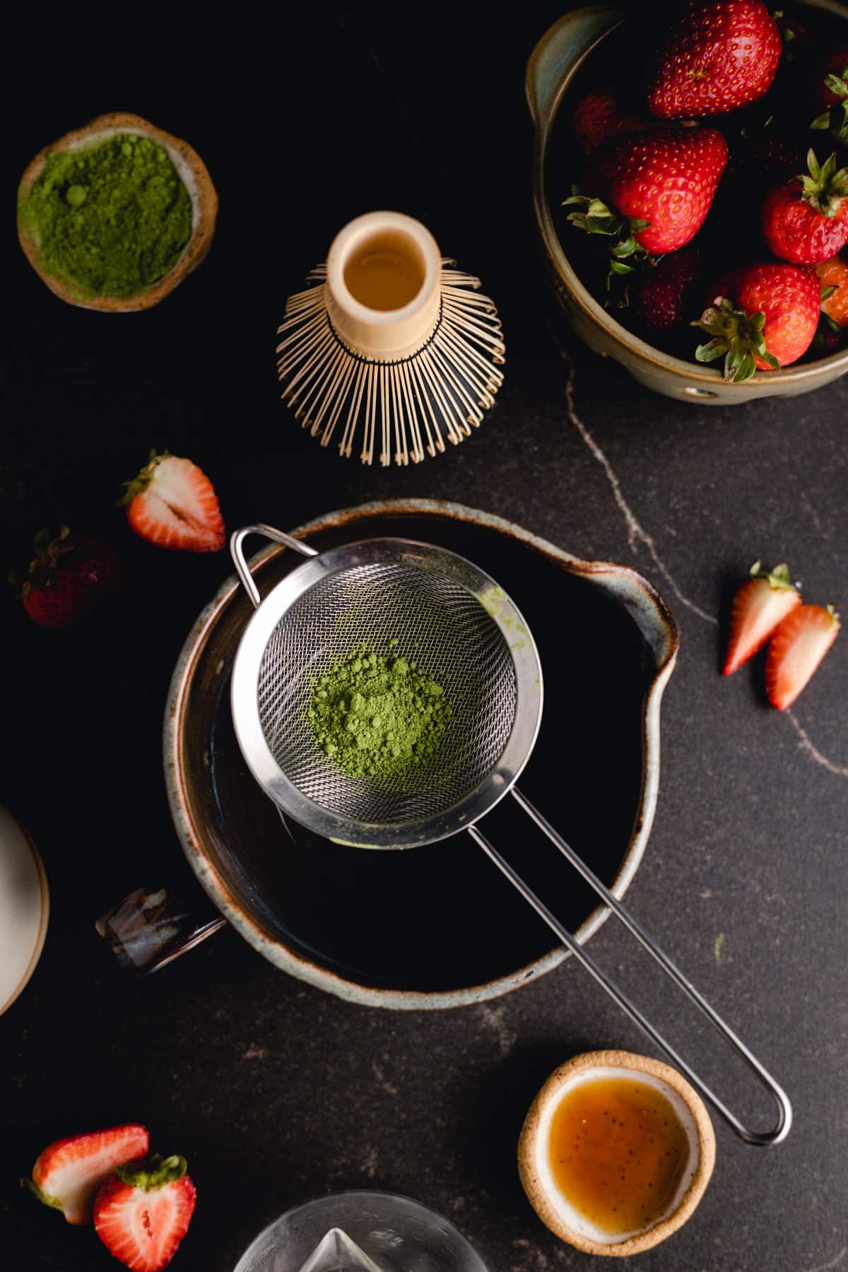 A sieve with matcha powder is placed over a ceramic pitcher. Surrounding items include strawberries, a bamboo whisk, a bowl of matcha powder, and a small bowl of honey on a dark surface.
