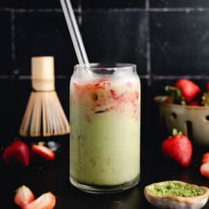 A glass of matcha latte mixed with strawberry syrup, ice cubes, and a clear straw, surrounded by strawberries, a bamboo whisk, a bowl of matcha powder, and a dark tiled background.