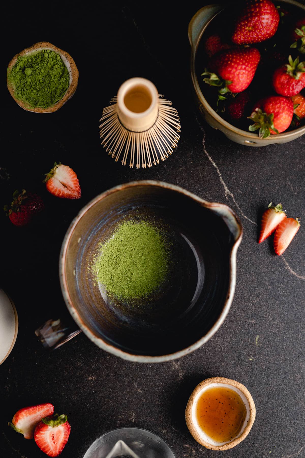 A ceramic bowl with green matcha powder, surrounded by a wooden whisk, fresh strawberries, a bowl of honey, and a bowl of extra matcha powder on a dark surface.