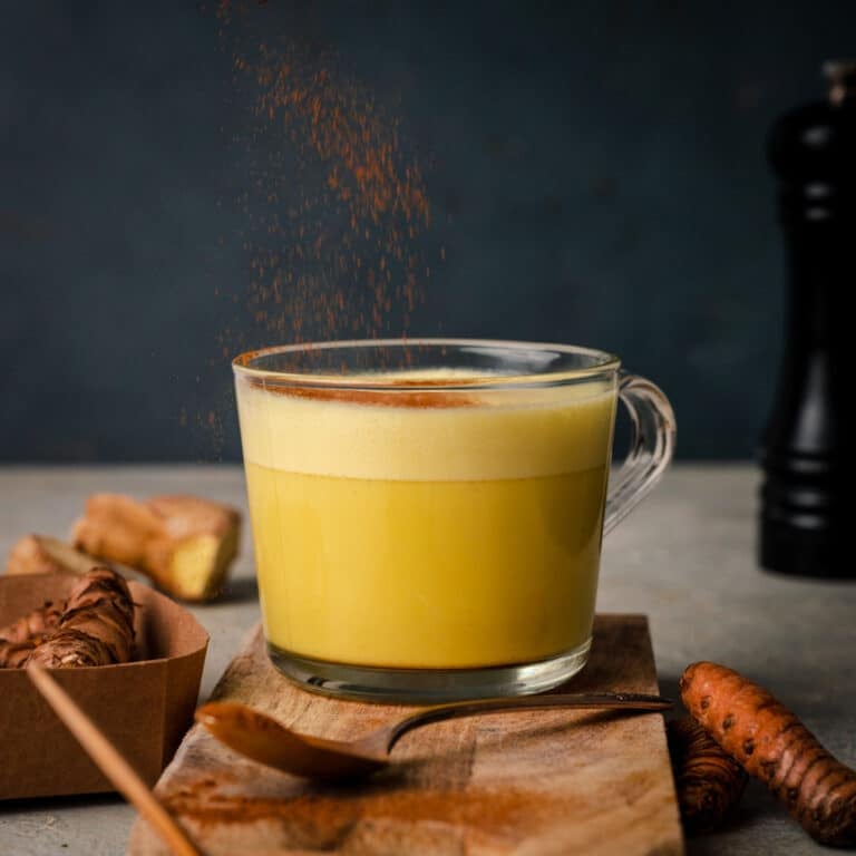A clear glass mug filled with yellow turmeric tea sits on a wooden board. Ground turmeric is sprinkled on top. A spoon rests nearby along with turmeric roots and a black pepper grinder in the background.