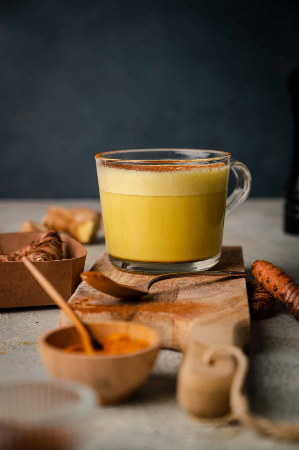A glass cup of yellow turmeric latte sits on a wooden board, surrounded by fresh turmeric roots and spices in small bowls.