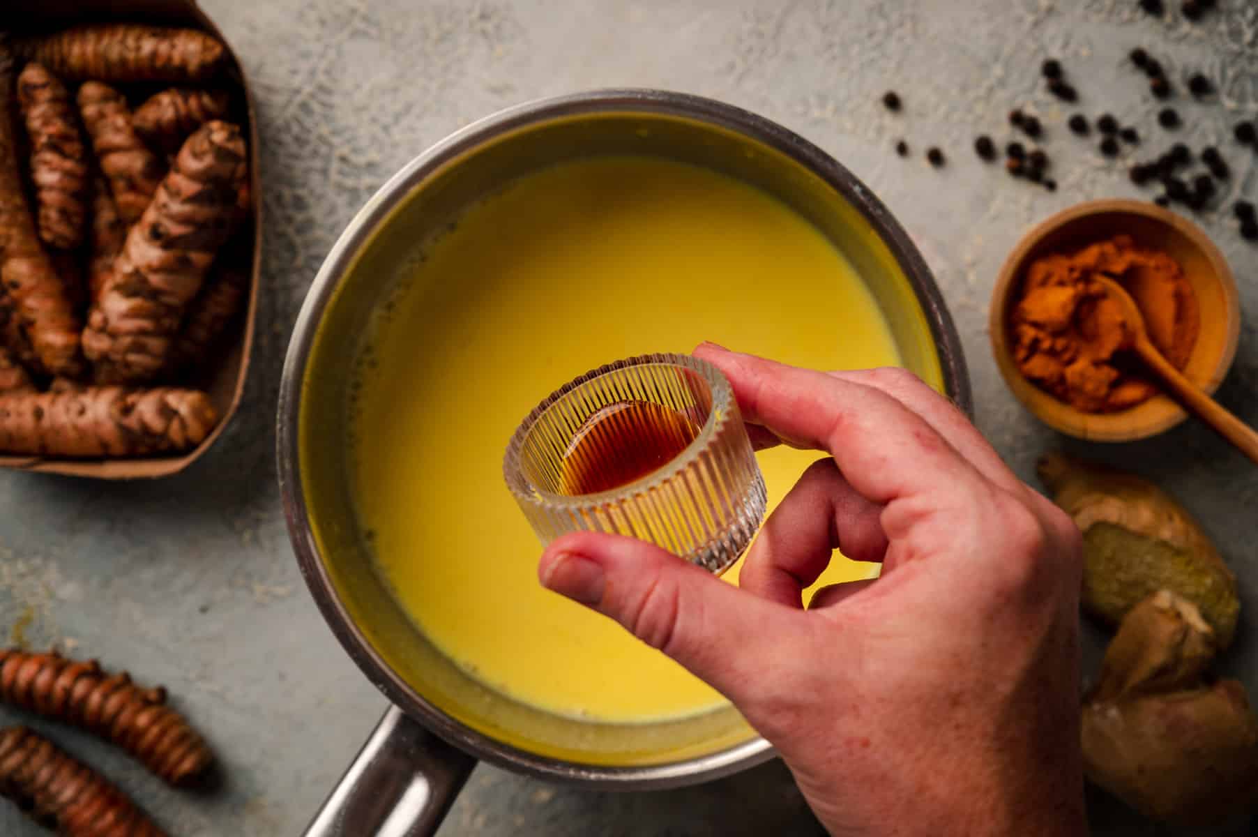 A hand adds an ingredient to a pot of yellow liquid, surrounded by bowls of spices and whole turmeric roots.
