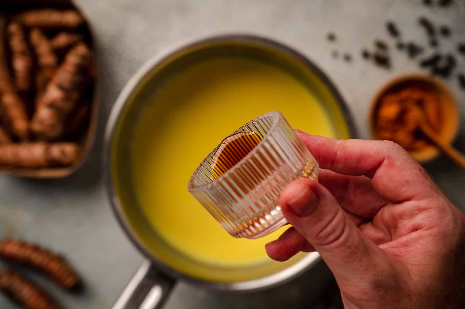 A hand holds a small glass container filled with a maple syrup substance above a saucepan containing a yellow liquid. Other ingredients are placed on the counter.