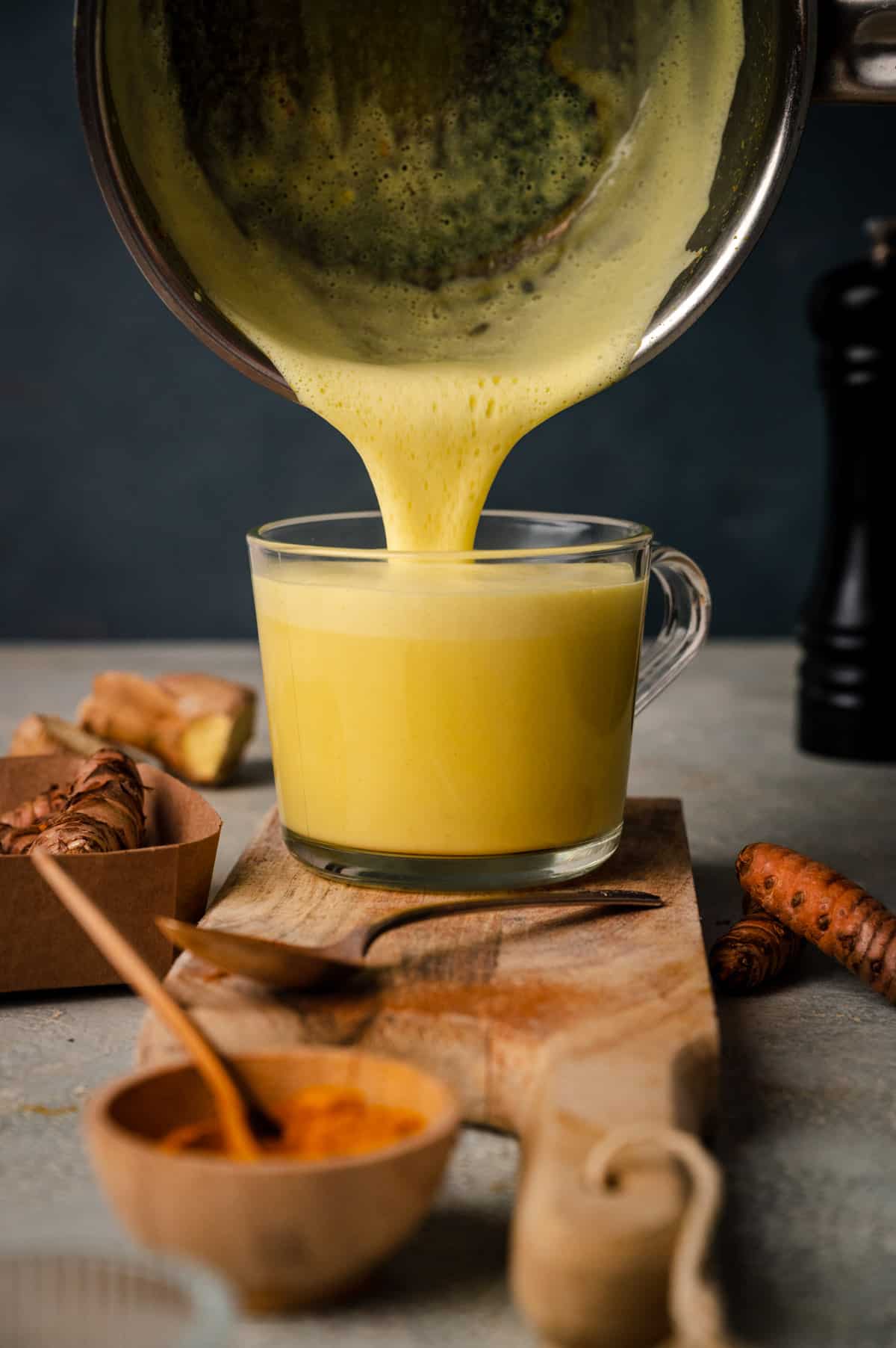A yellow beverage is being poured from a pot into a glass cup on a wooden board. Turmeric roots and powder in a bowl are visible in the background.