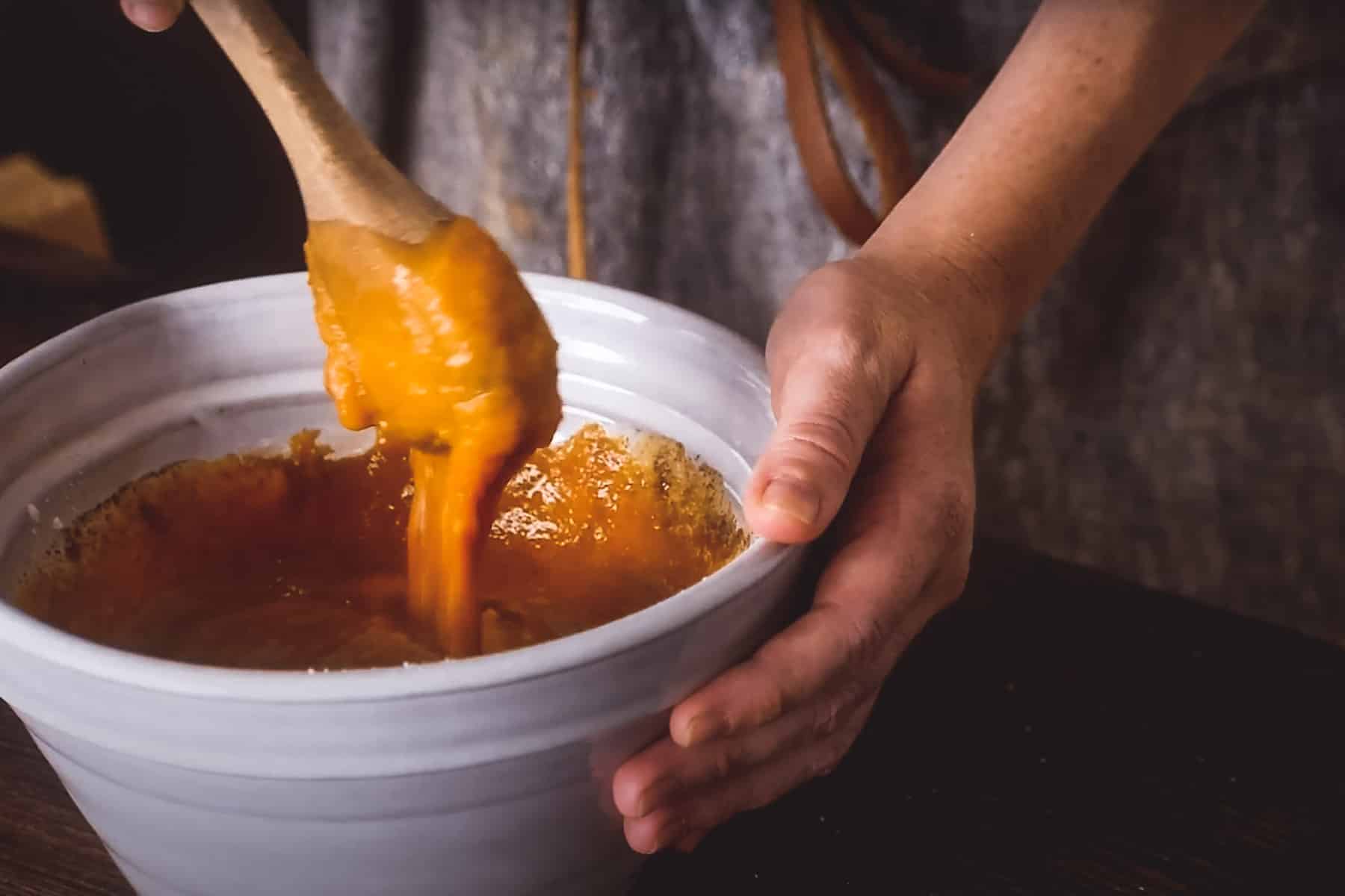 A person stirs a thick, pumpkin pie filling in a white bowl with a wooden spoon.