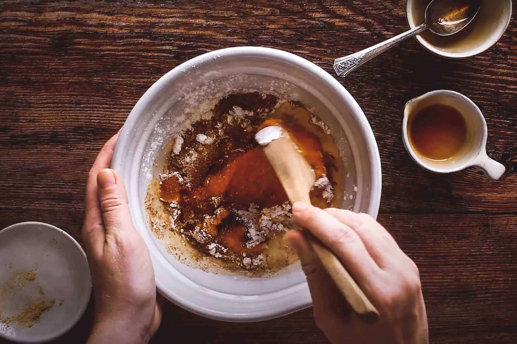 Hands stirring ingredients in a white bowl on a wooden surface, with a spoon.