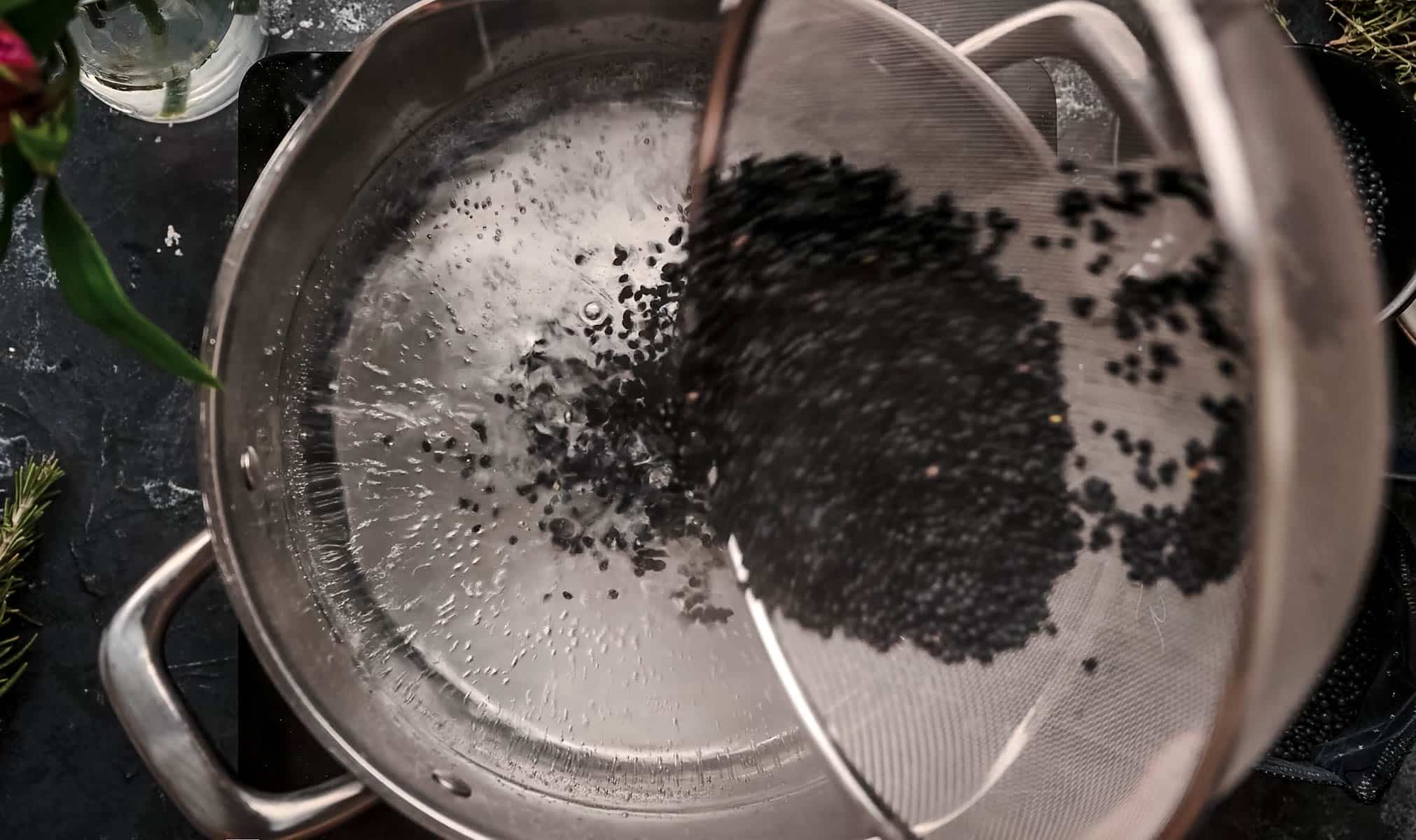 A sieve is used to pour black lentils into a stainless steel pot filled with water.