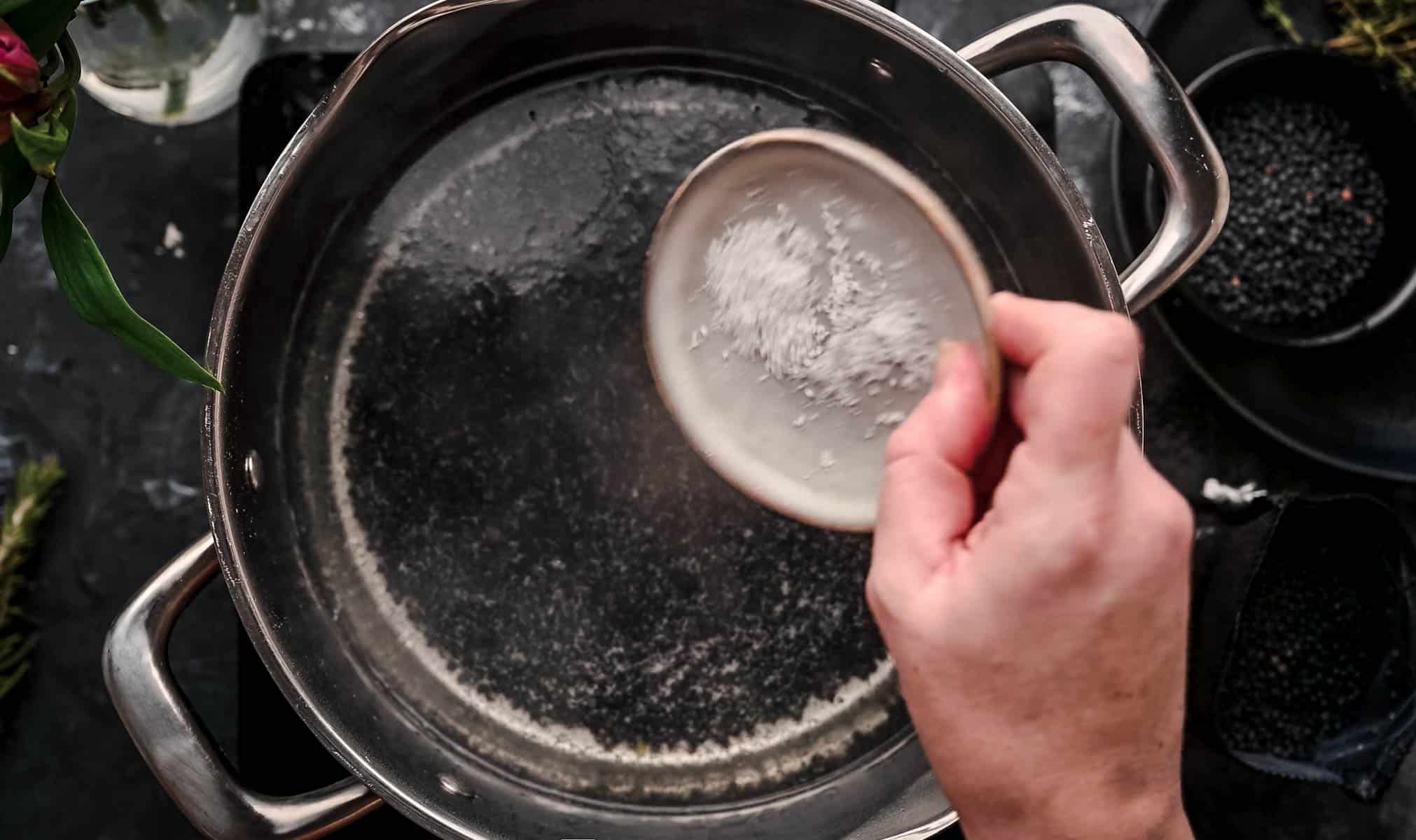 A hand is adding salt from a small dish into a pot of water on a stovetop.