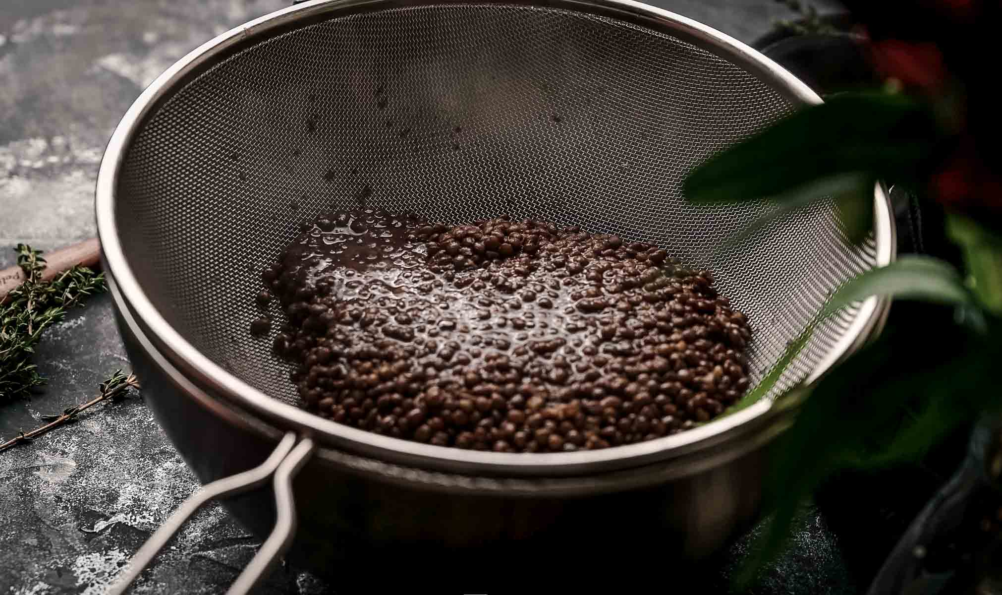 A close-up of lentils draining in a fine mesh sieve over a bowl on a dark surface, with some herbs visible in the background.