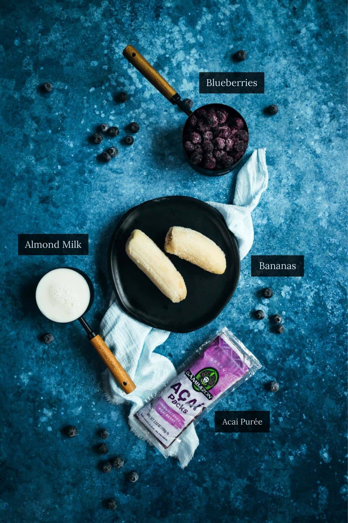 Top-down view of a black plate with two peeled bananas, a bowl of blueberries, a cup of almond milk, and a package of acai purée on a blue surface—perfect ingredients for crafting an irresistible vegan smoothie.
