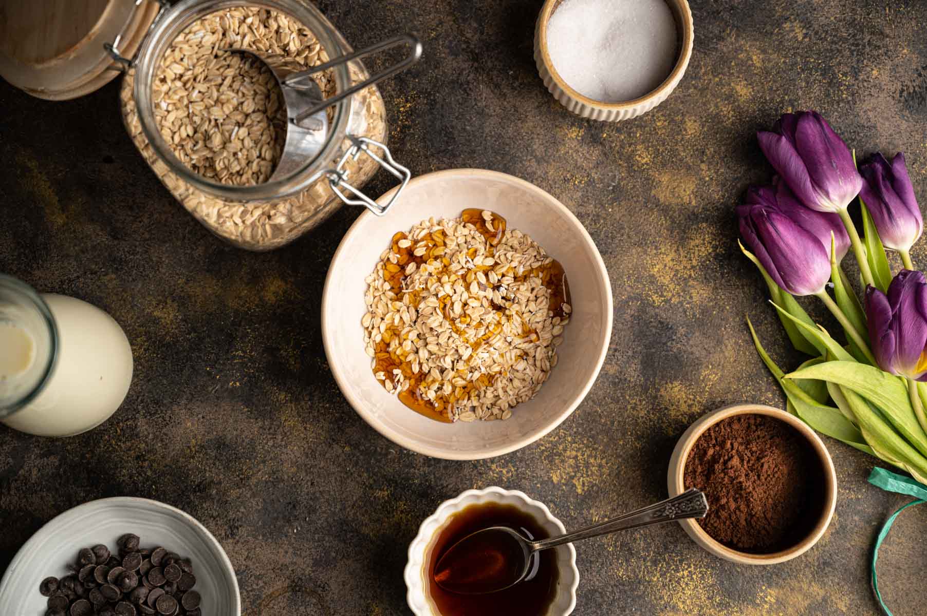 A bowl of oats with maple syrup surrounded by jars of oats, milk, chocolate chips, cocoa powder, and syrup.