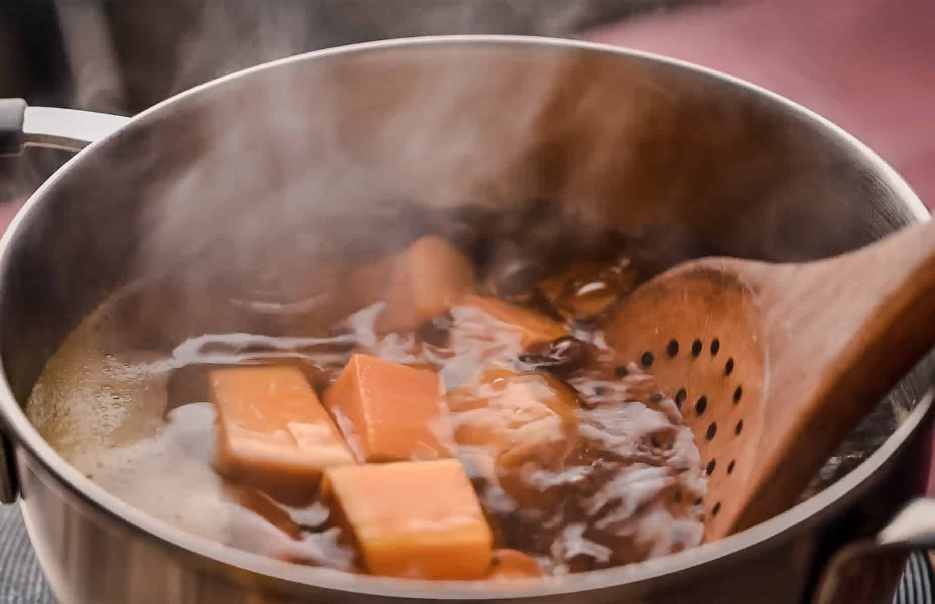 A pot of boiling water with chunks sweet potato and a slotted wooden spoon scooping them.