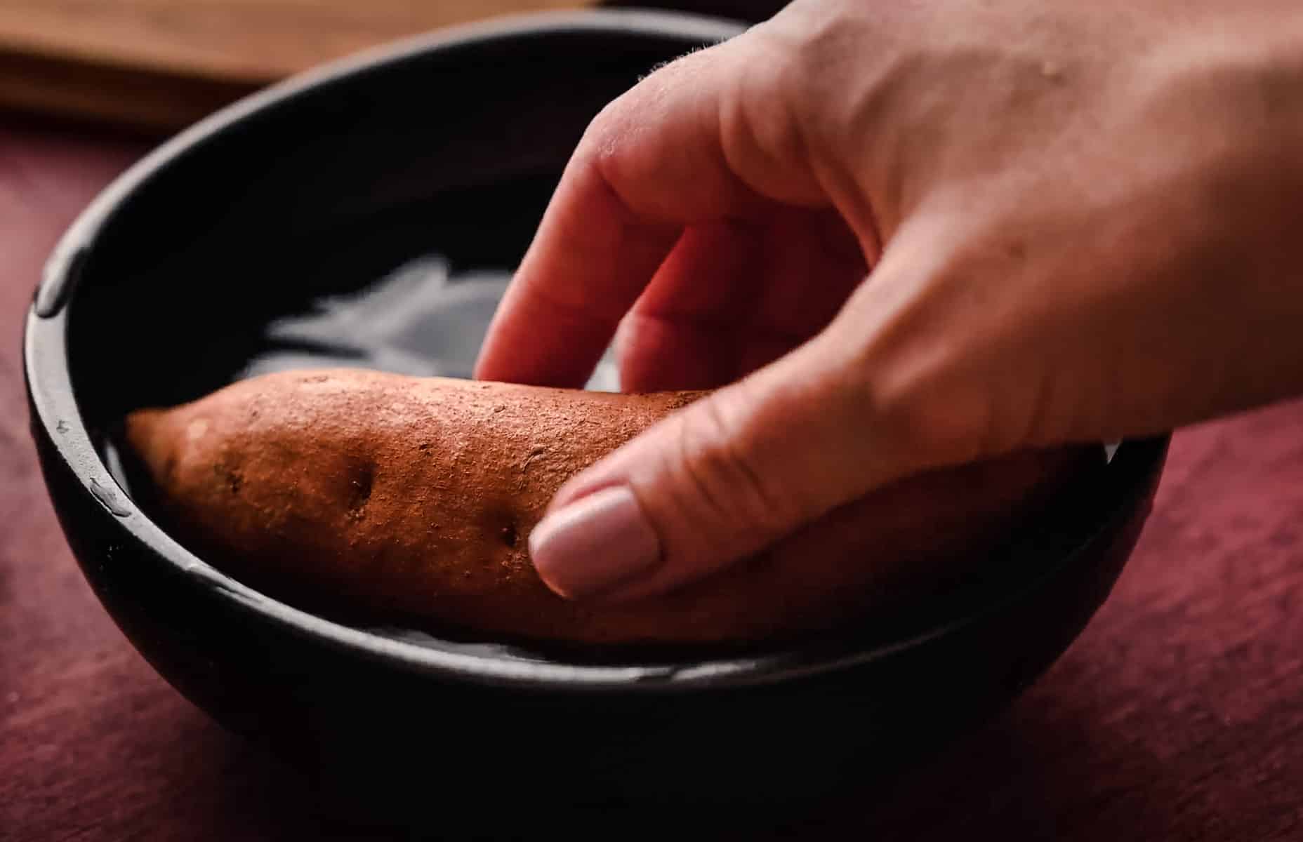A hand placing a raw sweet potato into a black bowl filled with water.