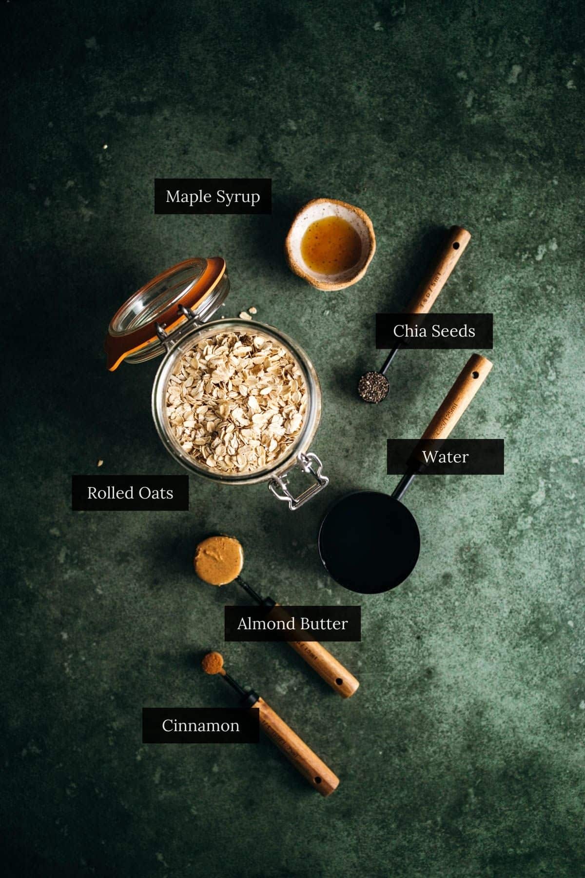 Overhead view of rolled oats, chia seeds, water, almond butter, cinnamon, and maple syrup on a dark surface.