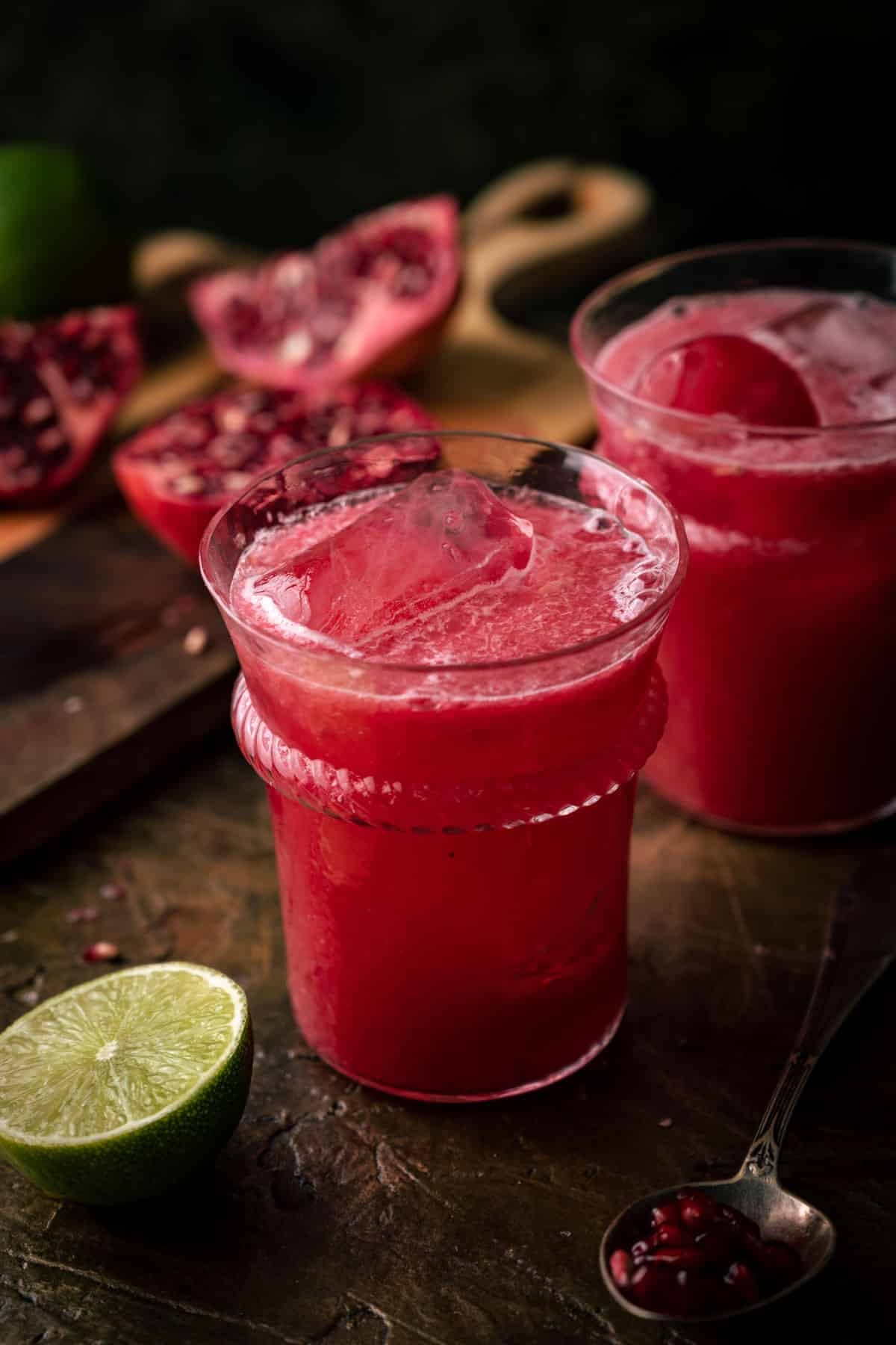 Two glasses of red pomegranate lime drink with ice are on a table.