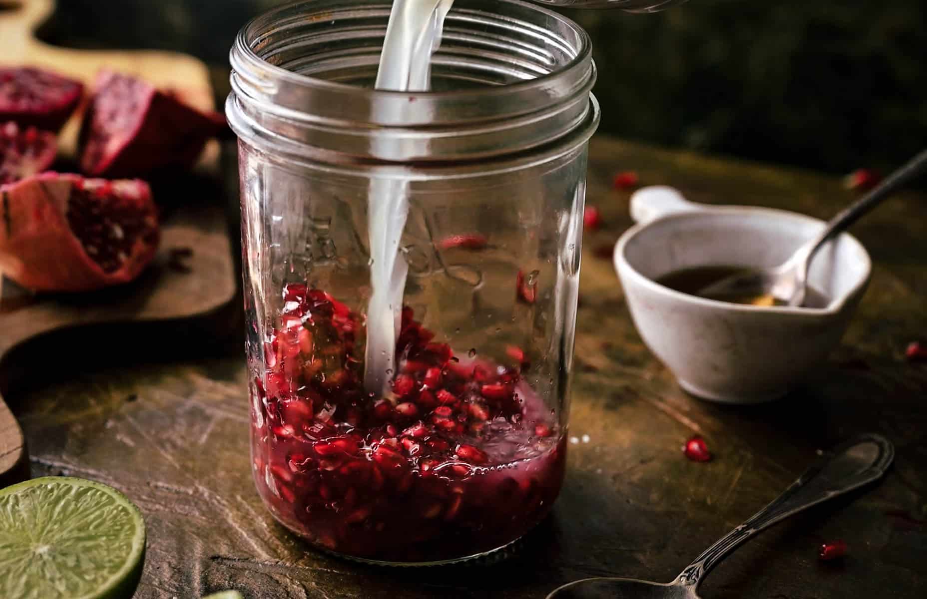 A glass jar being filled with a lime juice over pomegranate seeds.
