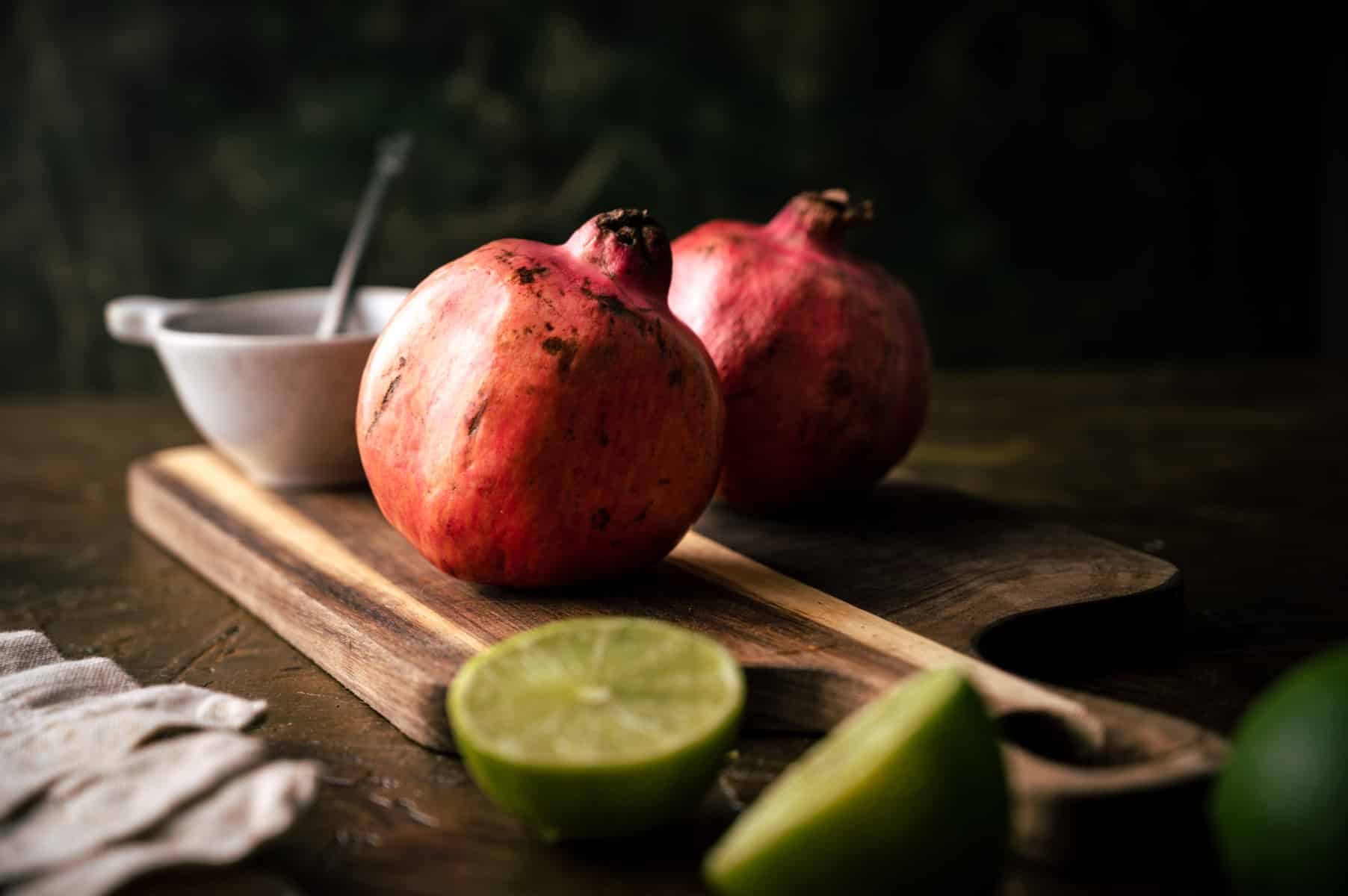 Two pomegranates rest on a wooden cutting board with halved limes and a white bowl.