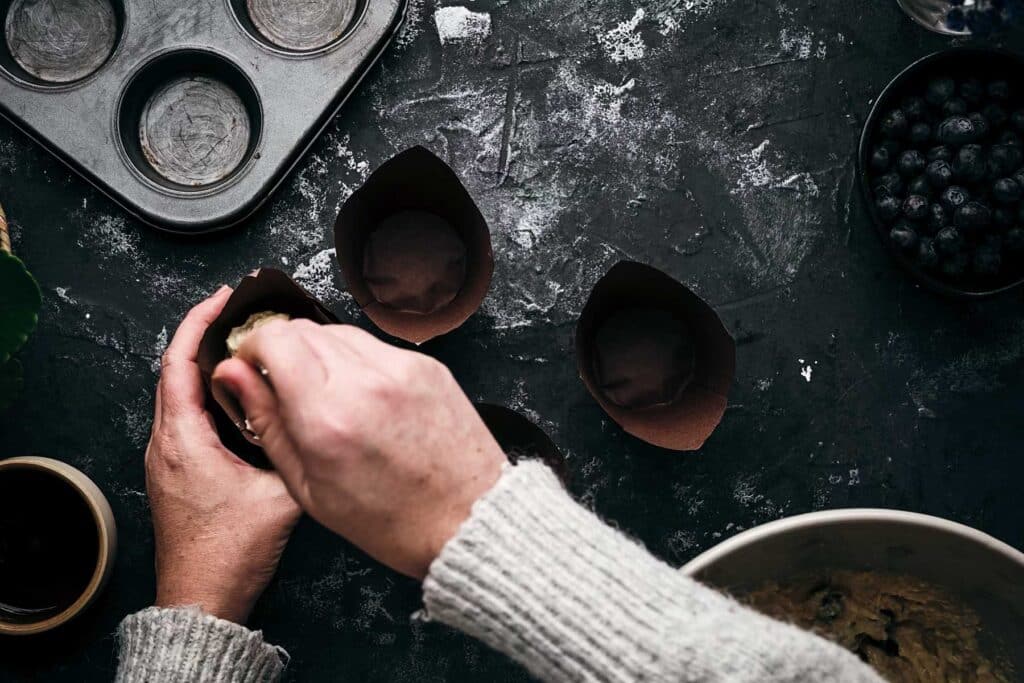 A person with a grey sweater is scooping muffin batter into paper liners.