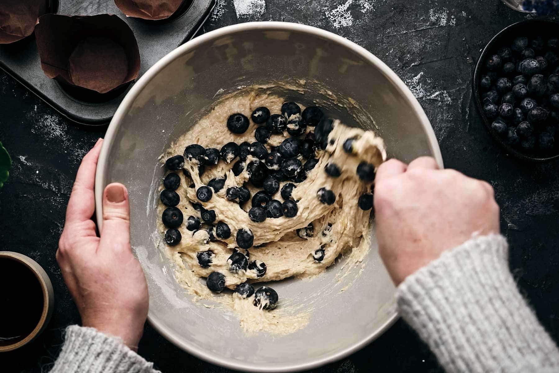 Hands mixing blueberry batter in a bowl, with a muffin tin and a bowl of blueberries nearby.