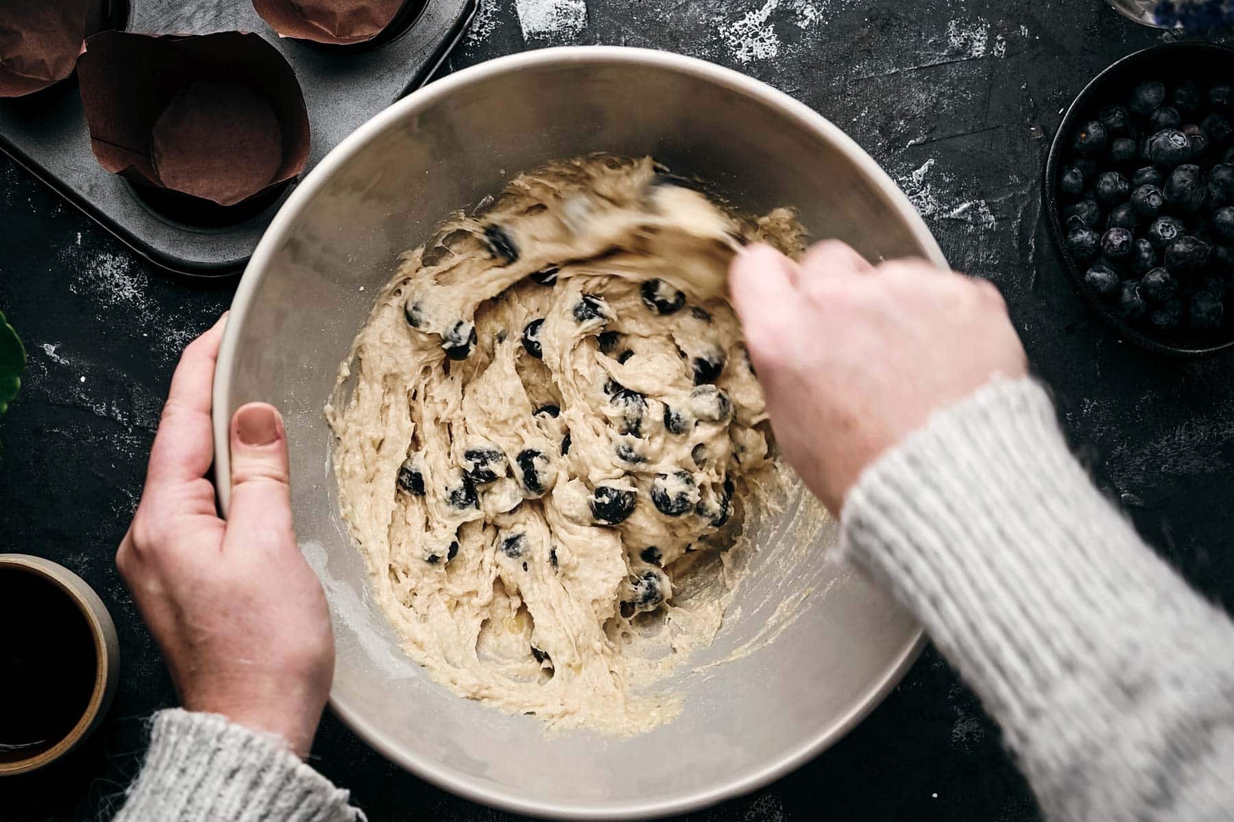 Hands stirring a bowl of batter with blueberries using a metal spoon.