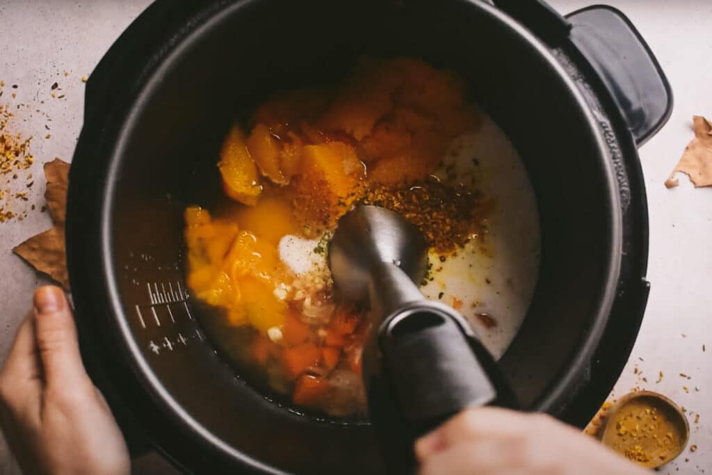 Hands using an immersion blender to blend an assortment of ingredients inside an electric pressure cooker.