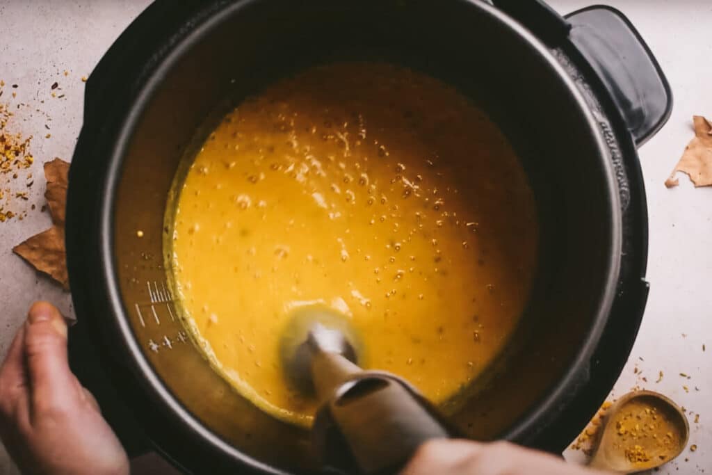 A person uses an immersion blender to blend a yellow soup in an Instant Pot.