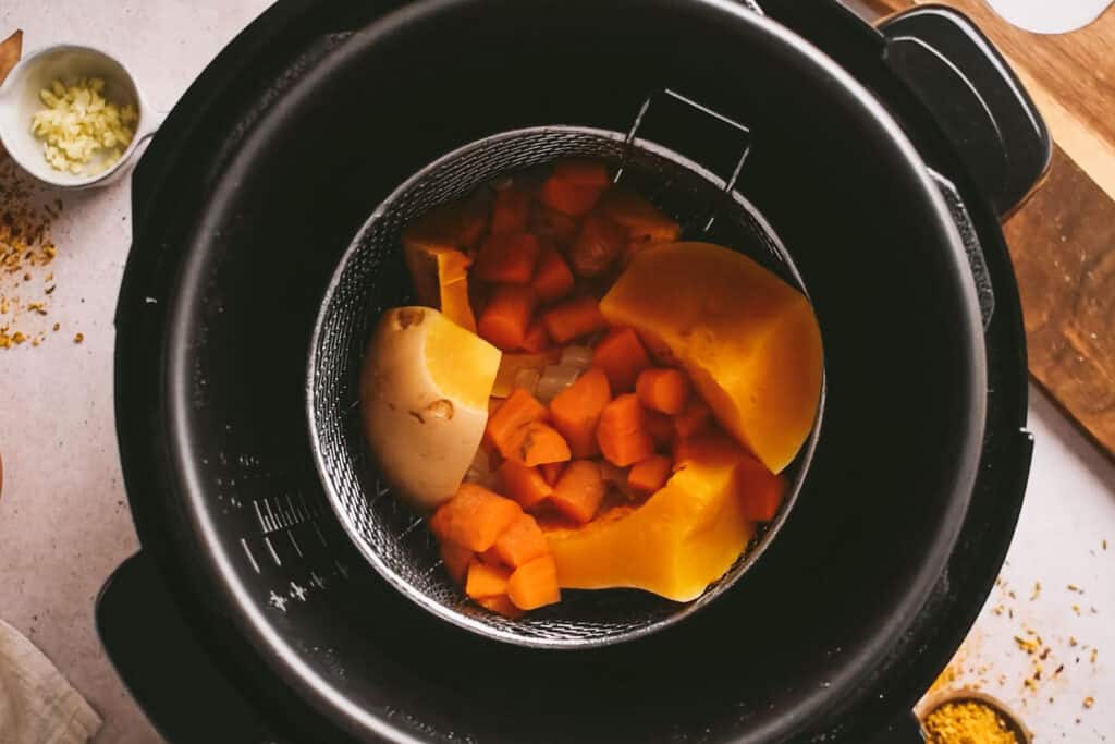 An overhead view of a pressure cooker with cooked pieces of squash and carrots inside the steamer basket.