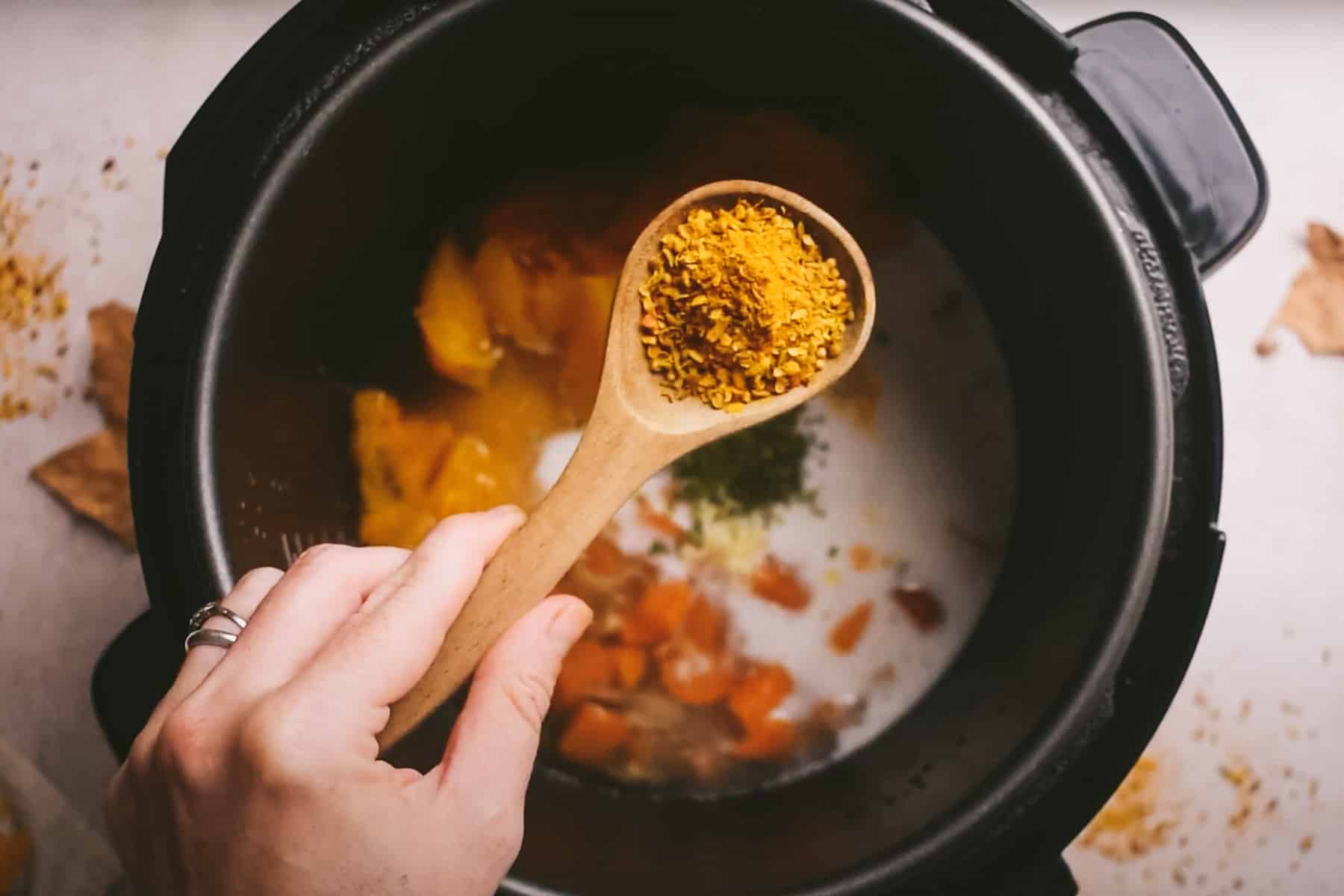 A hand holding a wooden spoon with curry powder over a black pot containing coconut milk, vegetables, and herbs.