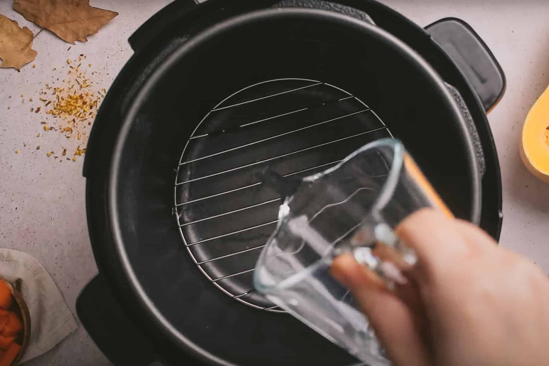 A hand is pouring water from a glass measuring cup into an empty pressure cooker.