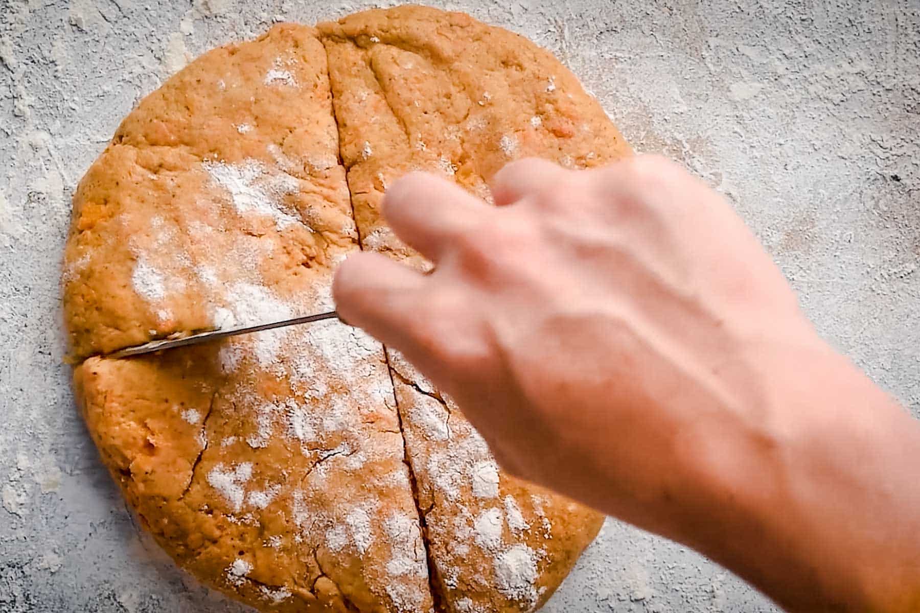 A person's hand is cutting a round, flour-dusted dough into sections with a knife on a lightly floured surface.