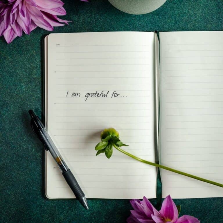 Open notebook with "I am grateful for..." written inside, lying beside a black pen and a green flower on a green surface, inviting you to develop an attitude of gratitude.