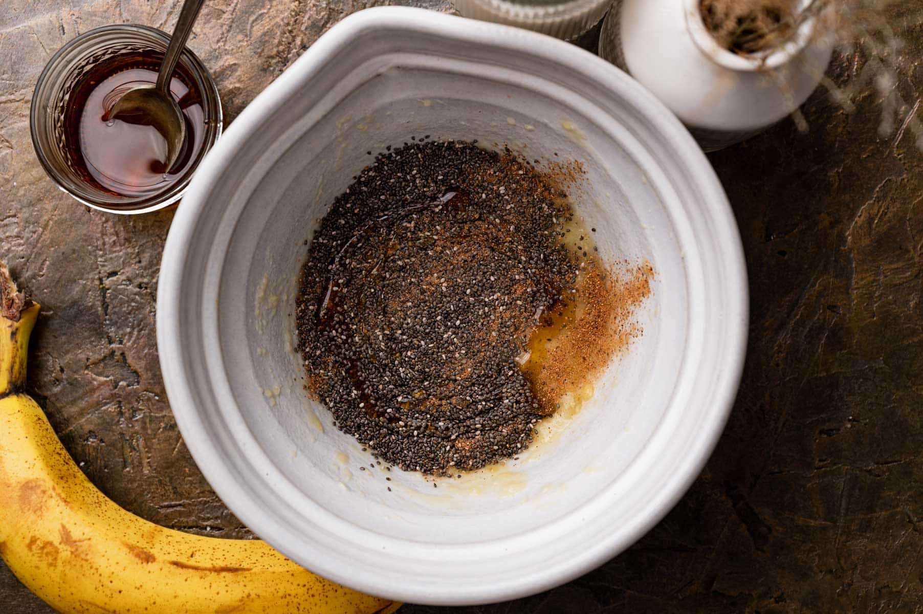 Bowl with chia seeds, spices, and liquid being mixed. A banana and a jar with a spoon are nearby on the table.