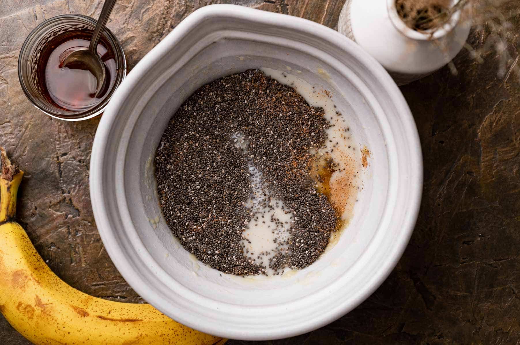 Bowl with chia seeds, milk, and spices on a table next to a banana and a jar of honey.