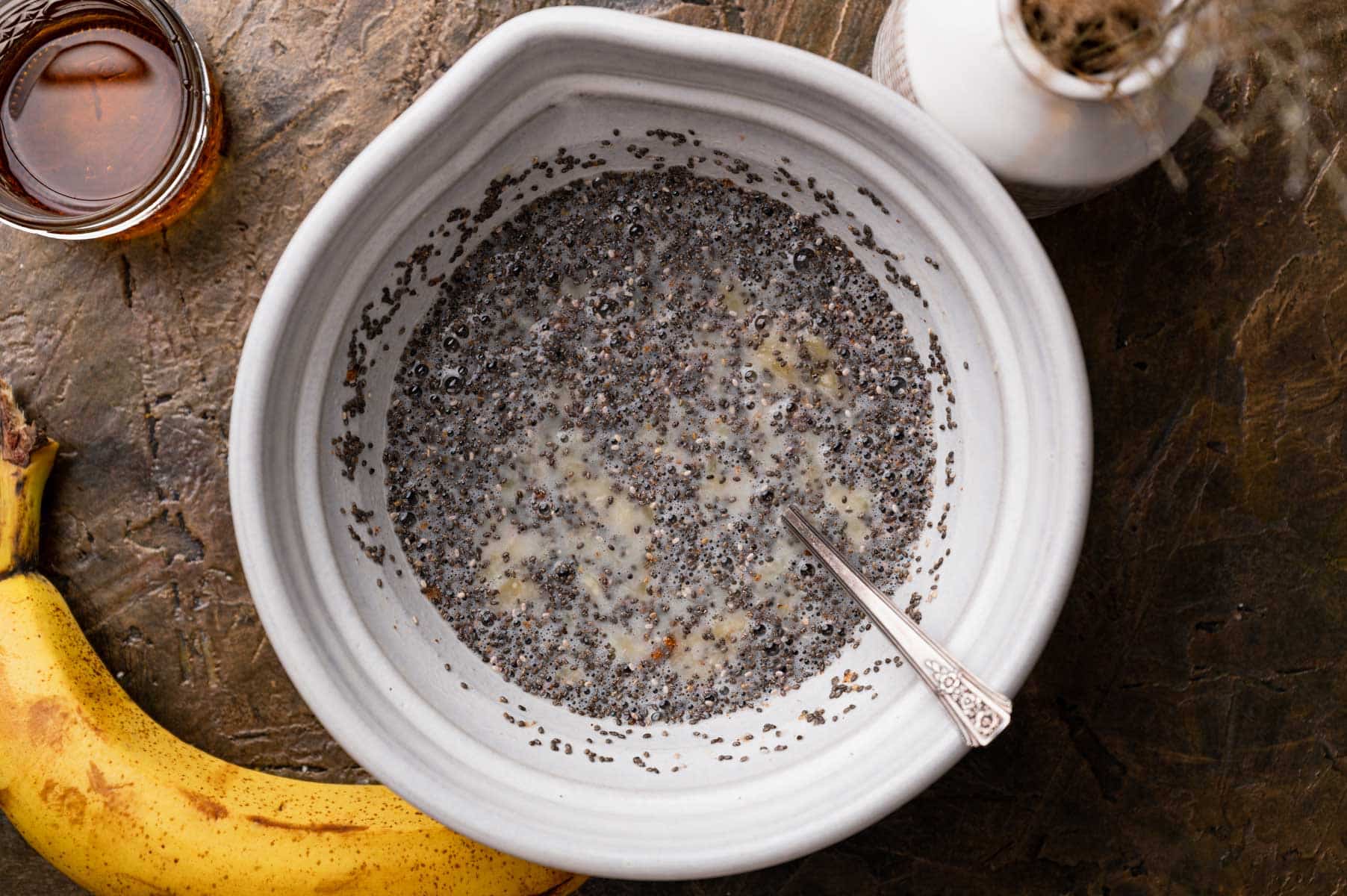 A ceramic bowl contains a mixture of chia seed pudding with a spoon. A banana and a small jar of maple syrup are nearby on a copper surface.