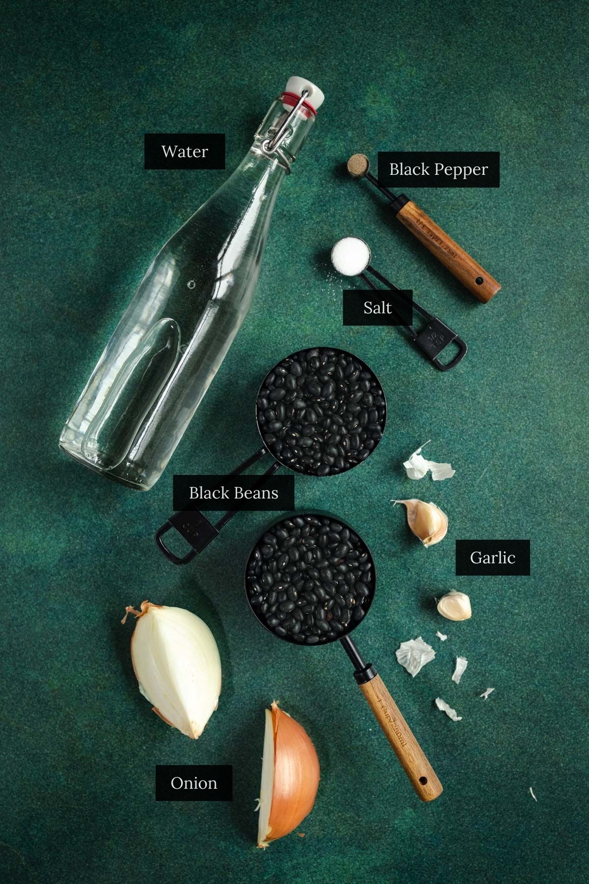 Ingredients for cooking black beans: water, black beans, black pepper, salt, garlic cloves, and a halved onion on a green background.