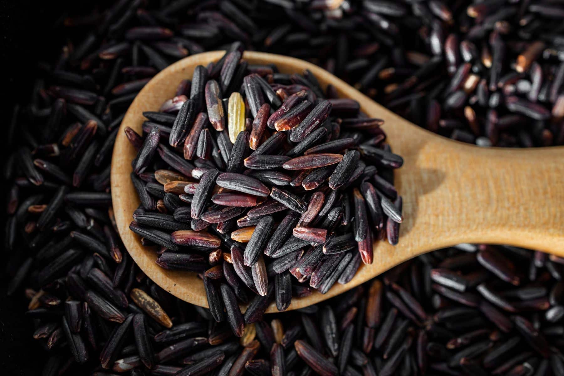 A wooden spoon holds a pile of uncooked black rice grains with more grains scattered underneath.