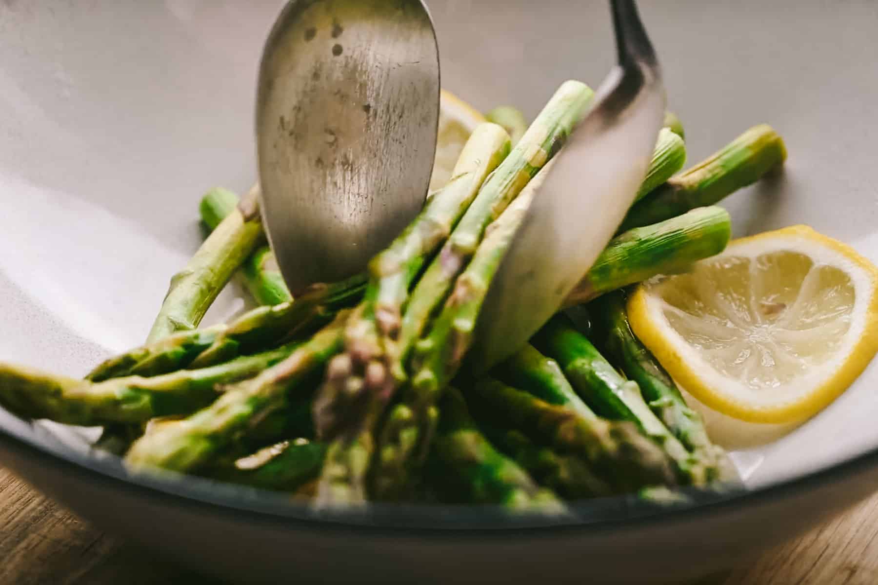 A bowl containing steamed asparagus spears, garnished with lemon slices, with two metal serving utensils.