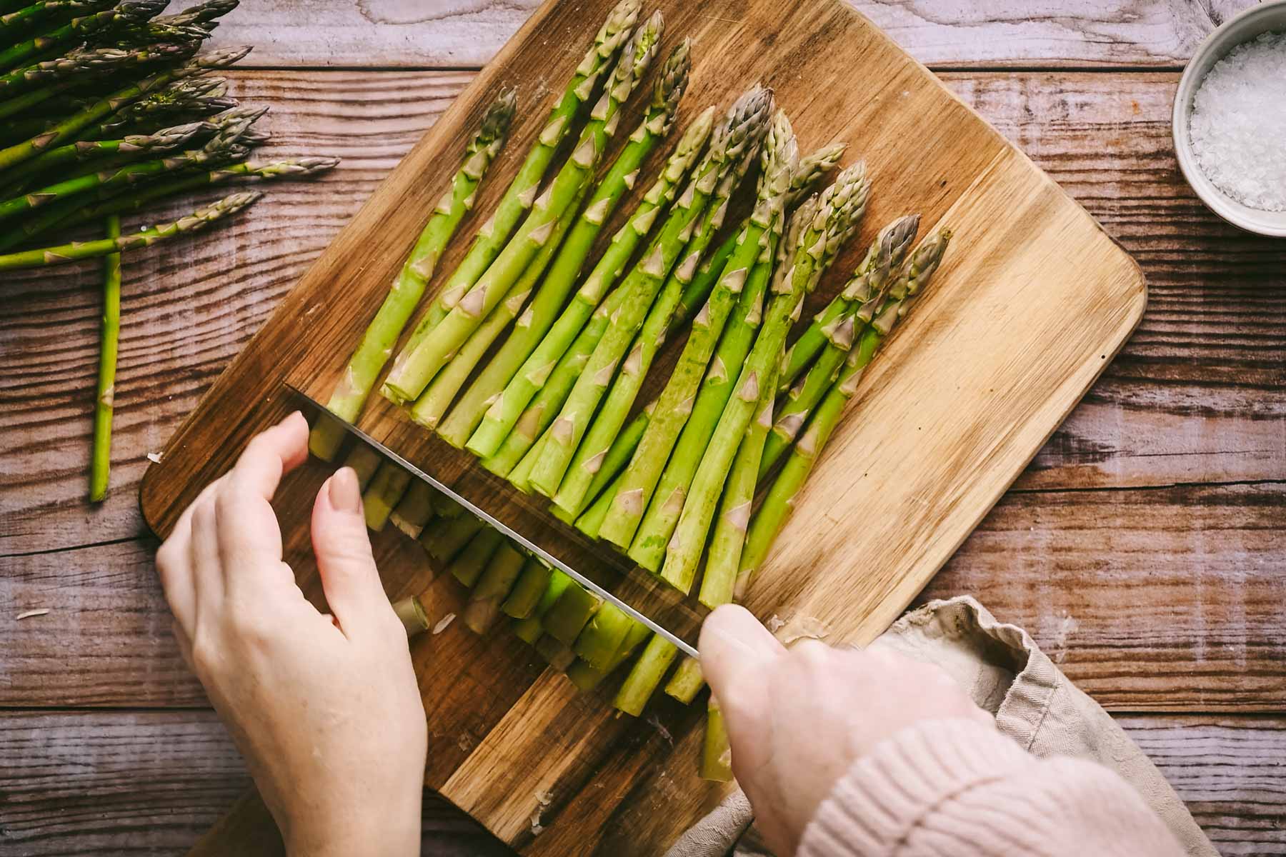 Hands cutting fresh asparagus on a wooden board with a knife, with a bowl of salt nearby.
