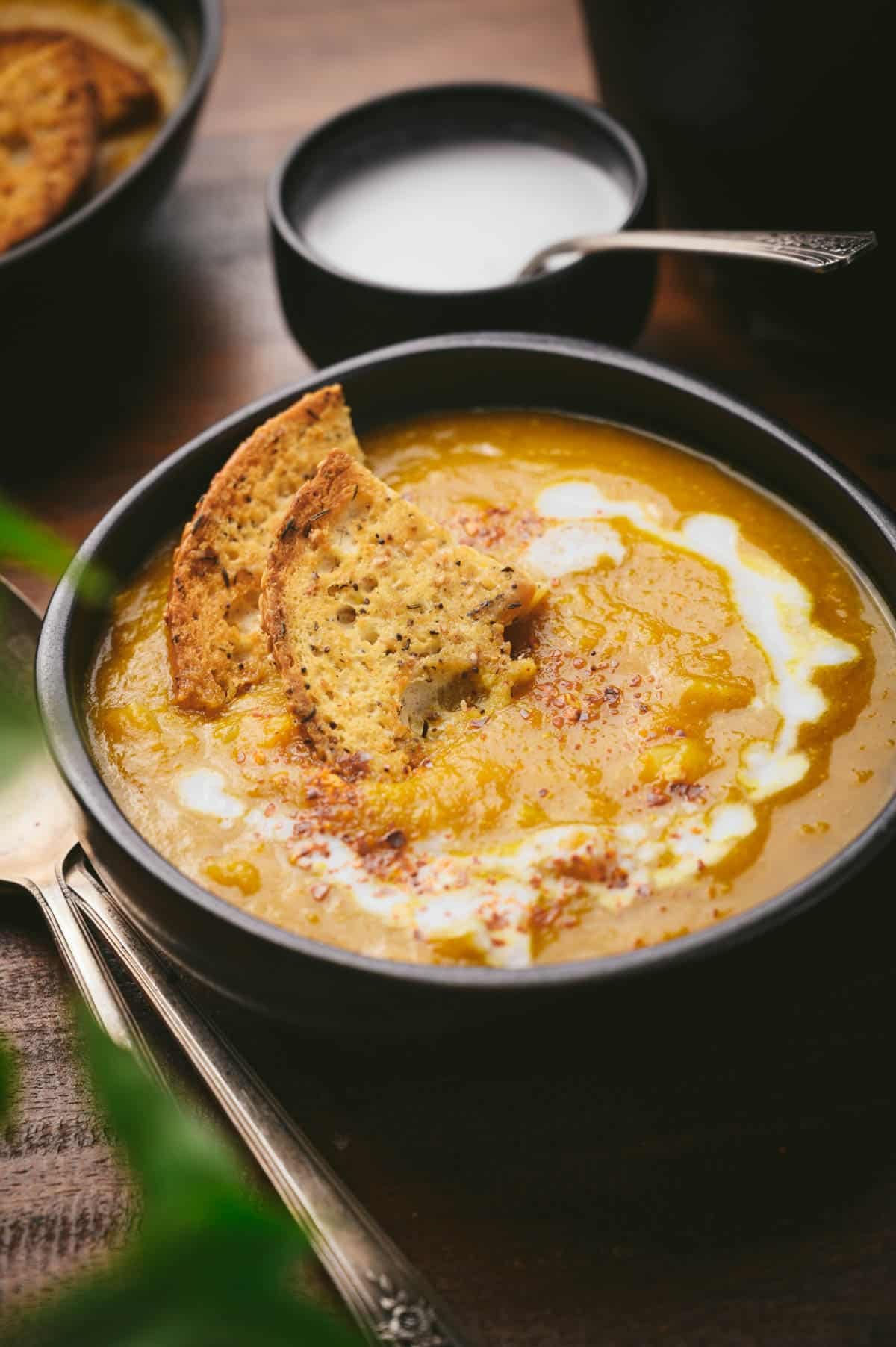 A bowl of soup with coconut cream swirls, topped with crispy bread slices. A spoon rests beside it.